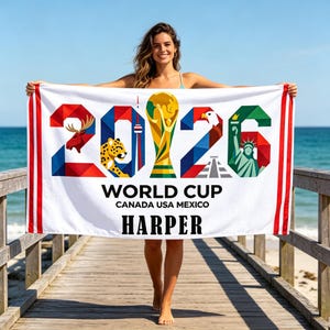 May include: A white rectangular beach towel featuring the text 2026 WORLD CUP CANADA USA MEXICO and the name HARPER. The design includes a colorful geometric graphic of the World Cup trophy, a moose, a jaguar, a tower, and the Statue of Liberty.