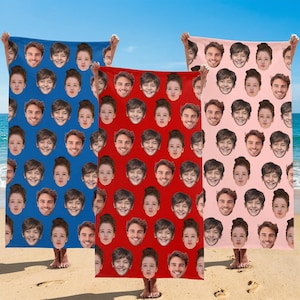May include: Three beach towels are displayed, each held by a person. One is blue, one is red, and one is pink. Each towel features a repeating pattern of faces. The towels are on a sandy beach with the sea in the background.