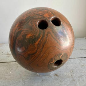 May include: A brown and orange bowling ball with three finger holes. The ball has a marbled pattern and is resting on a clear stand. The ball is designed for the sport of bowling.