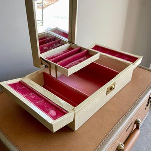 May include: A vintage jewelry box with a cream-colored exterior and a hinged mirror. The interior features compartments lined with pink velvet. The box is open, revealing multiple storage levels. It sits atop a brown suitcase.