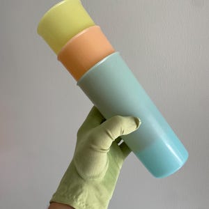 May include: Three pastel-colored, cylindrical containers stacked together. The top container is yellow, the middle one is peach, and the bottom one is light blue. The containers are held by a green glove.
