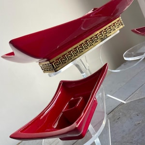 May include: Red, glossy, decorative serving dishes with a gold Greek key design on a clear stand. The dishes have a unique, angular shape and are displayed on a clear, tiered stand.