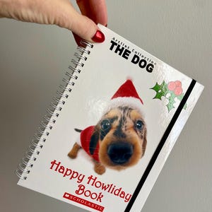 May include: A spiral-bound book with a festive design. The cover features a dachshund wearing a Santa hat and a red sweater. Text includes "Artist Collection THE DOG" and "Happy Howliday Book" in red. A Scholastic logo is at the bottom.