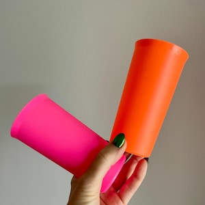 May include: Two vibrant, neon-colored plastic tumblers. One is bright pink, the other is a vivid orange. The tumblers have a cylindrical shape with a slightly flared rim. The background is a plain, light gray.