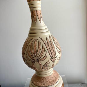 May include: Cream-colored table lamp with a textured, floral design in shades of peach and brown. The lamp has a rounded base and a long, slender neck. The lamp's shade is a light yellow color.