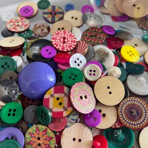 May include: A collection of various buttons in different colors, shapes, and sizes. The assortment includes wooden, plastic, and fabric-covered buttons, with a mix of solid colors, patterns, and textures. A large purple sphere is also present.