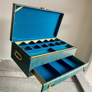 May include: A teal and gold jewelry box with a hinged lid and a pull-out drawer. The box has a patterned design and gold accents.
