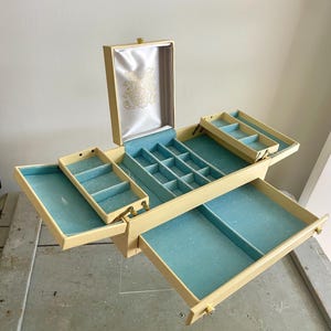 May include: A cream-colored jewelry box with an open lid featuring a white satin lining with a gold floral design. The interior is lined with light blue velvet and includes multiple tiered compartments, a central grid section, and a pull-out drawer.