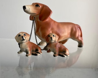 May include: A set of three ceramic dachshund figurines. The largest dachshund is brown and white with a chain around its neck. The two smaller dachshunds are brown and white and are standing next to the larger dachshund.