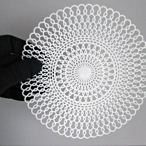 May include: A white, circular, crocheted doily with a delicate, intricate pattern. The doily is made of a fine, white thread and has a circular design with a central point.