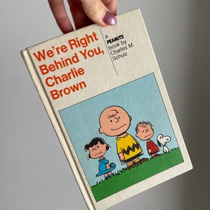 May include: A vintage book titled "We're Right Behind You, Charlie Brown" by Charles M. Schulz. The cover features Charlie Brown, Lucy, Linus, and Snoopy on a green grassy area with a blue sky background.