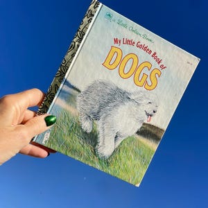 May include: A vintage "Little Golden Book" titled "My Little Golden Book of DOGS." The cover features a fluffy white dog running through a grassy field. The book has a blue sky background.