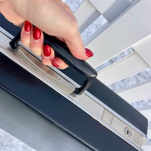 May include: Black Samsonite briefcase with silver trim and a black handle. The briefcase has a lock and the brand name is visible on the silver trim. The case is being held by a person.