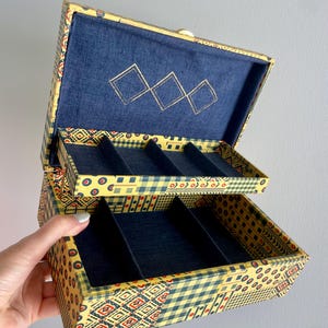 May include: Open jewelry box with a yellow, red, and blue patterned exterior. The interior is lined with dark blue fabric and features multiple compartments for storage. The box is open, revealing the interior and the compartments.