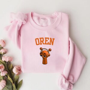 May include: A light pink sweatshirt with the name "OREN" in orange block letters. Below the text is a cartoon character wearing headphones. The character is orange with brown accents.