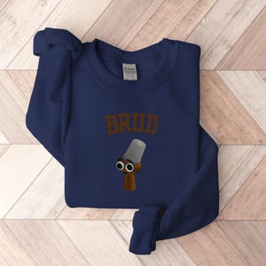 Puede incluir: Sudadera azul marino con la palabra "BRUD" en letras marrones. Debajo del texto hay una ilustraci&oacute;n de dibujos animados de un objeto marr&oacute;n con una parte superior gris y dos ojos negros.