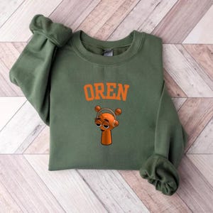 May include: An olive green sweatshirt with the name "OREN" in orange block letters. Below the text is a cartoon character with headphones. The character is orange with a long neck and a small head.