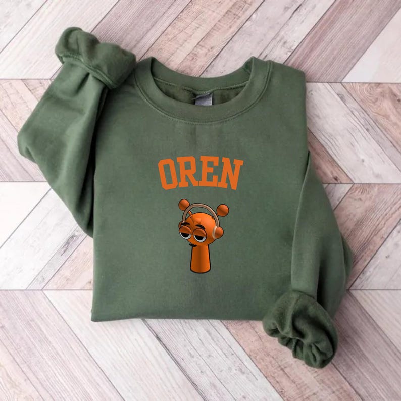 May include: An olive green sweatshirt with the name "OREN" in orange block letters. Below the text is a cartoon character with headphones. The character is orange with a long neck and a small head.