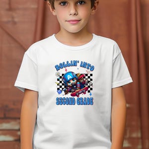 May include: White t-shirt with a graphic of a cartoon character on a skateboard, wearing a helmet with the letter "A". The text "ROLLIN' INTO SECOND GRADE" is in blue. The design includes a checkered background.