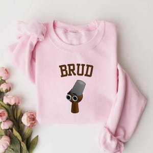 Puede incluir: Sudadera rosa claro con la palabra "BRUD" en letras marrones. Debajo del texto hay una ilustraci&oacute;n de dibujos animados de una figura marr&oacute;n con un sombrero gris y dos ojos grandes y redondos.