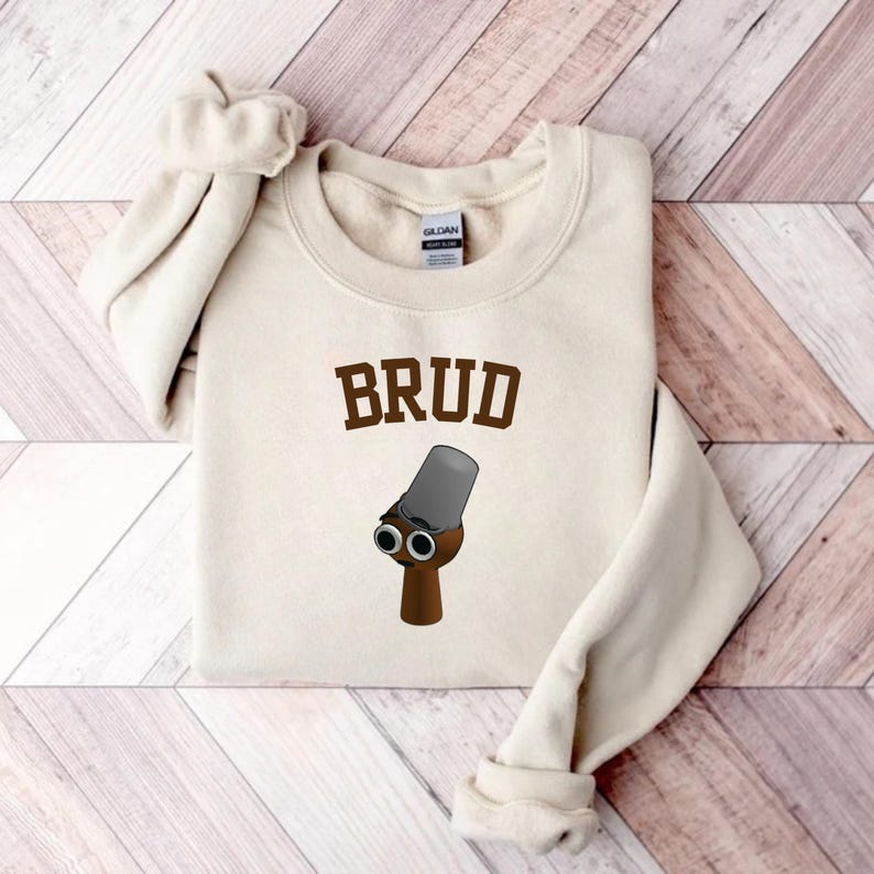 Puede incluir: Una sudadera color crema con la palabra "BRUD" en letras marrones. Debajo del texto hay una ilustraci&oacute;n de dibujos animados de una figura marr&oacute;n con un cubo gris como cabeza y dos ojos negros.