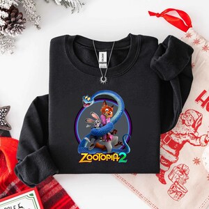 May include: Black sweatshirt featuring a colorful graphic of characters from Zootopia 2, including a fox, a rabbit, and a blue snake. The text "Zootopia 2" is displayed in a stylized font below the characters. A silver necklace is visible.