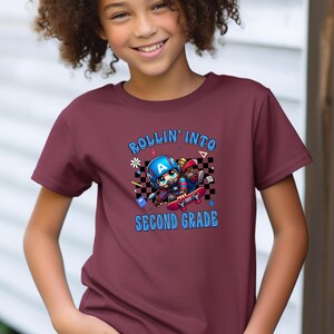 May include: Burgundy t-shirt with the text "ROLLIN' INTO SECOND GRADE" in blue. The shirt features a cartoon graphic of a character on a skateboard, wearing a helmet and superhero mask. The design includes a checkered background and stars.