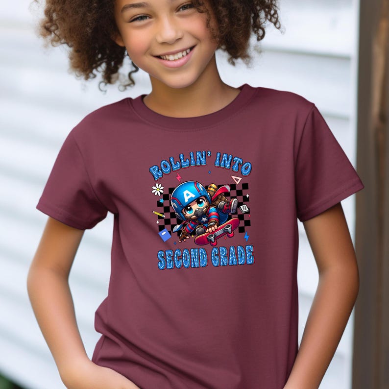 May include: Burgundy t-shirt with the text "ROLLIN' INTO SECOND GRADE" in blue. The shirt features a cartoon graphic of a character on a skateboard, wearing a helmet and superhero mask. The design includes a checkered background and stars.