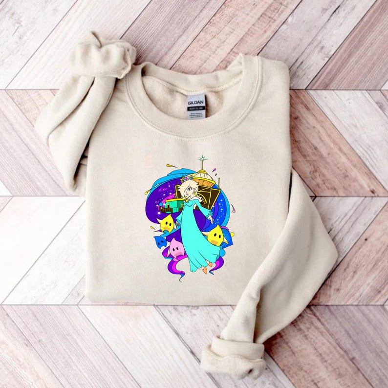 May include: A beige sweatshirt featuring a colorful graphic of a character in a blue dress, surrounded by stars and a space-themed background. The design includes yellow stars and purple accents.