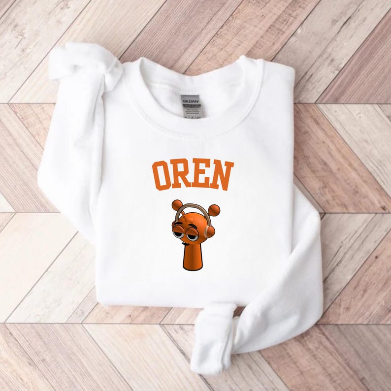May include: White sweatshirt with the word "OREN" in orange block letters above a cartoon character wearing headphones. The character is orange with brown eyes and a small body. The sweatshirt is on a light wood grain surface.