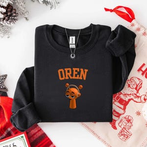 May include: Black crew neck sweatshirt with the name "OREN" in orange block letters above a cartoon character wearing headphones. The character is orange with brown eyes and a small body. A silver necklace is visible.