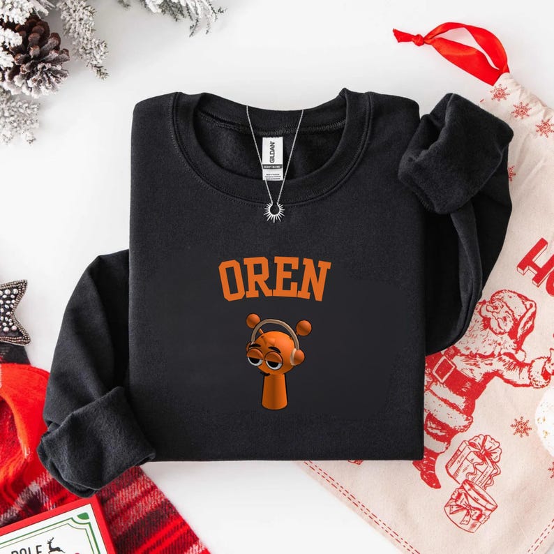 May include: Black crew neck sweatshirt with the name "OREN" in orange block letters above a cartoon character wearing headphones. The character is orange with brown eyes and a small body. A silver necklace is visible.