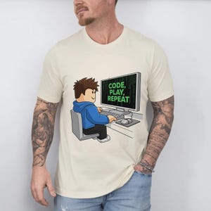 May include: Cream-colored t-shirt featuring a cartoon illustration of a person coding on a computer. The computer screen displays the text "CODE, PLAY, REPEAT" in green. The person is wearing a blue hoodie. The design is centered on the shirt.