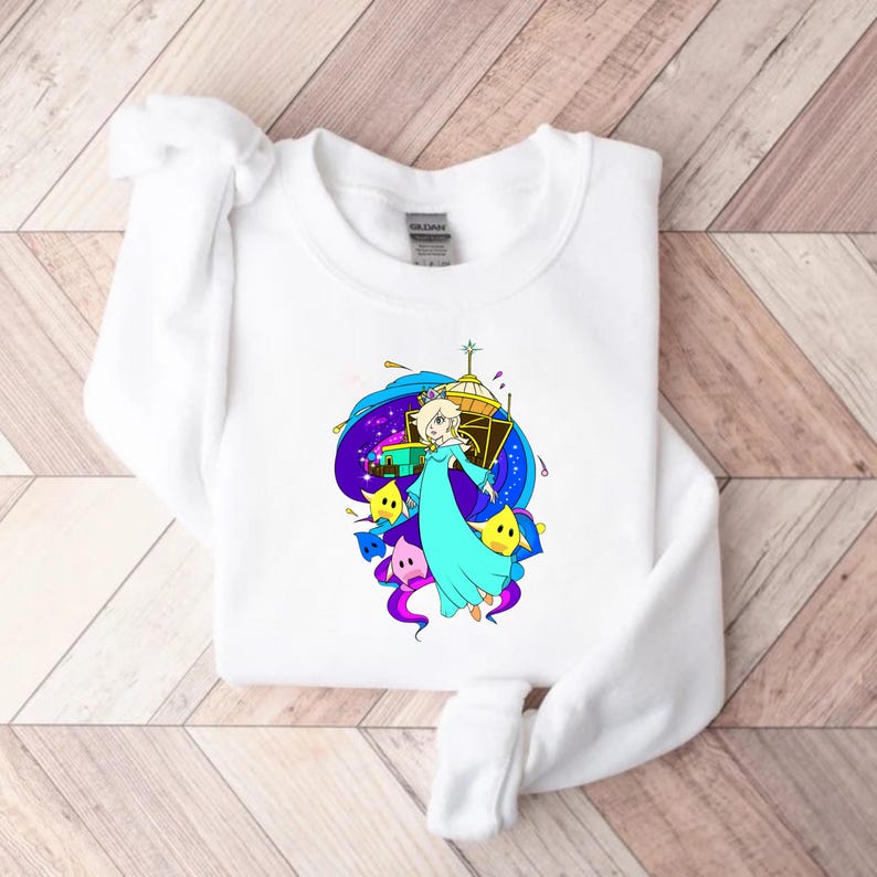 May include: White sweatshirt featuring a colorful graphic of a character in a blue dress with a crown, surrounded by stars and a space-themed background. The design includes yellow, purple, and blue hues, creating a vibrant and playful aesthetic.