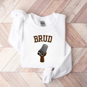 Puede incluir: Sudadera blanca con la palabra "BRUD" en letras marrones. Debajo, una ilustraci&oacute;n de dibujos animados de un objeto marr&oacute;n con dos ojos redondos y un cilindro gris encima.