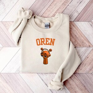 May include: Cream-colored sweatshirt with the word "OREN" in orange block letters. Below the text is a cartoon character with orange skin, wearing headphones. The sweatshirt is folded on a wooden surface.