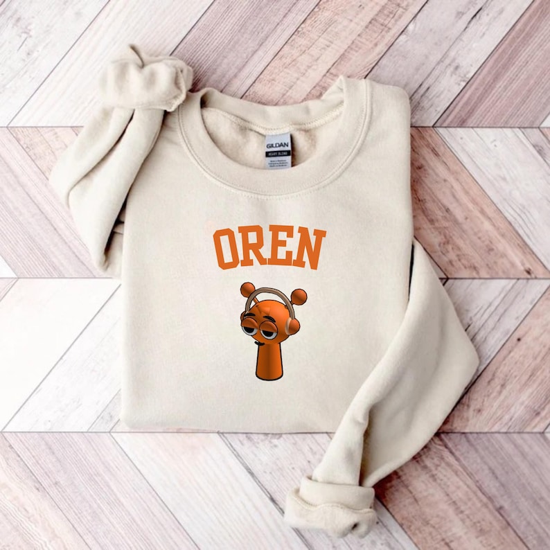 May include: Cream-colored sweatshirt with the word "OREN" in orange block letters. Below the text is a cartoon character with orange skin, wearing headphones. The sweatshirt is folded on a wooden surface.