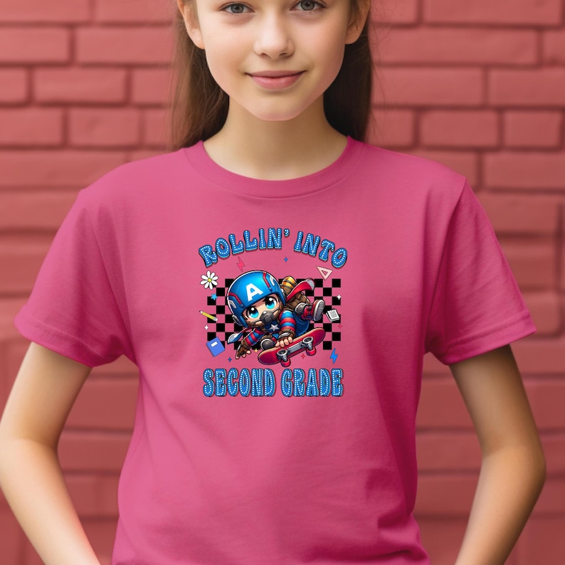 May include: A pink t-shirt featuring a graphic of a cartoon character on a skateboard, wearing a blue helmet with the letter "A". The text "ROLLIN' INTO SECOND GRADE" is above and below the graphic. The shirt is being worn by a person.