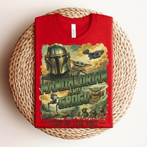Puede incluir: Una camiseta roja con un estampado gr&aacute;fico de The Mandalorian y Grogu. El dise&ntilde;o incluye el texto "The Mandalorian and Grogu" y "This is the way" junto con ilustraciones de un personaje con casco, peque&ntilde;as criaturas verdes y una nave espacial.