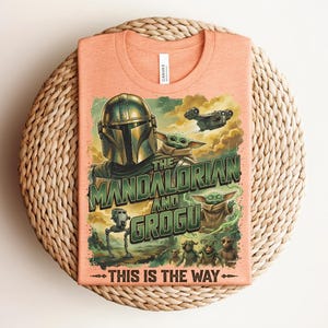 Puede incluir: Una camiseta color melocot&oacute;n con un estampado gr&aacute;fico de The Mandalorian y Grogu. El dise&ntilde;o incluye el texto "THE MANDALORIAN AND GROGU" y "THIS IS THE WAY" junto con ilustraciones de un personaje con casco, peque&ntilde;as criaturas verdes y una nave espacial.