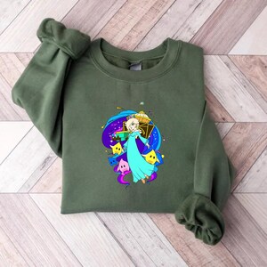 May include: An olive green sweatshirt featuring a colorful graphic of a character in a blue dress, surrounded by stars and a cosmic background. The design includes yellow and purple stars, and a gold crown.