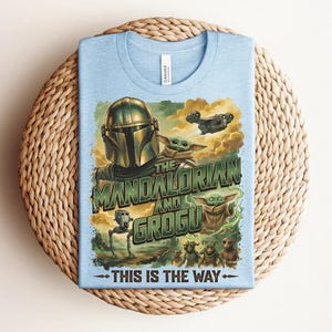 Puede incluir: Una camiseta azul claro con un estampado gr&aacute;fico de The Mandalorian y Grogu. El dise&ntilde;o incluye el texto "THE MANDALORIAN AND GROGU" y "THIS IS THE WAY" junto con ilustraciones de un personaje con casco, una peque&ntilde;a criatura verde y una nave espacial.