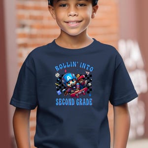 May include: Navy blue t-shirt with a graphic of a cartoon character wearing a Captain America helmet on a skateboard. The text "ROLLIN' INTO SECOND GRADE" is in blue and white.
