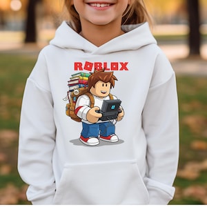 May include: White hoodie featuring a Roblox graphic. The graphic shows a cartoon character with a backpack full of books and a handheld gaming device. The word "ROBLOX" is printed in red above the character.