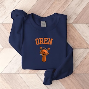 May include: Navy blue sweatshirt with the word "OREN" in orange block letters. Below the text is a cartoon character with orange skin, headphones, and large eyes. The sweatshirt is made of a soft material.