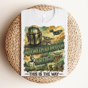 Puede incluir: Una camiseta blanca con un estampado gr&aacute;fico de The Mandalorian y Grogu. El dise&ntilde;o incluye el texto "THE MANDALORIAN AND GROGU" y "THIS IS THE WAY" junto con ilustraciones de un personaje con casco, peque&ntilde;as criaturas verdes y una nave espacial.