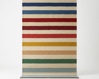 May include: A rectangular area rug featuring a repeating pattern of horizontal stripes in navy blue, light blue, deep red, burgundy, mustard yellow, tan, and forest green, all separated by wide bands of cream-colored fabric throughout the design.