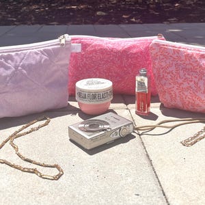 May include: An assortment of beauty and fashion accessories, including three cosmetic bags, makeup, jewelry, and a vintage camera. The bags are pink and white, with one quilted and two patterned. A gold chain and bracelet are also visible.