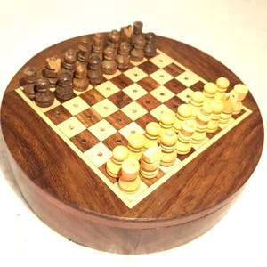 May include: A round wooden chess set with a chessboard inlaid in the center. The chess pieces are arranged on the board, with dark brown and light beige pieces. The set is made of dark brown wood with a light wood border.