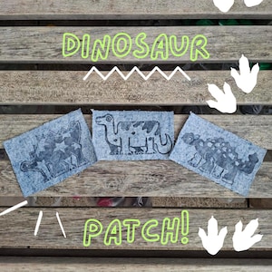 May include: Three rectangular denim fabric patches featuring dark blue screen-printed dinosaur illustrations. The left patch shows a stegosaurus with stars, the centre a long-necked dinosaur, and the right an ankylosaurus. Text reads DINOSAUR PATCH!